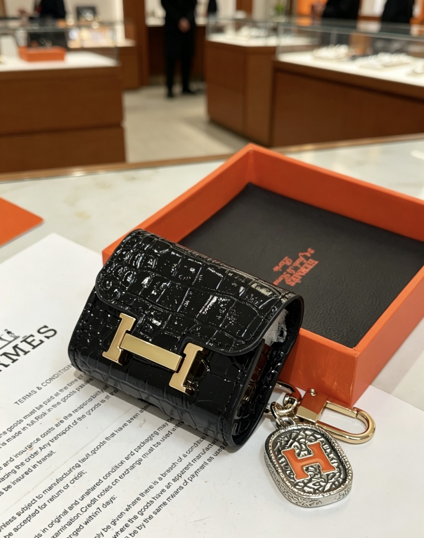 Hermes Constance AirPods Case Bag Charm pendentif Cuir verni noir embossé crocodile Quincaillerie en métal doré Charme en métal argenté avec émail orange Détail H Taille unique pour le charme AirPods standard longueur totale environ 8 cm